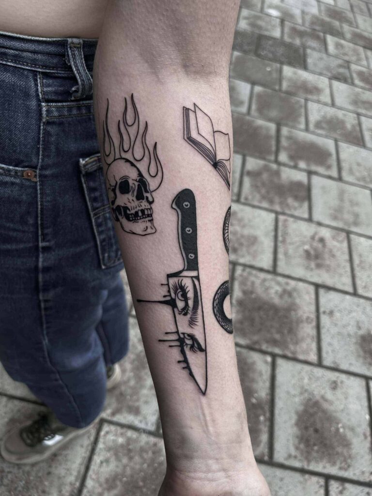 Blackwork tattoos on a forearm of a knife with eyes, a burning skull and a opened book.