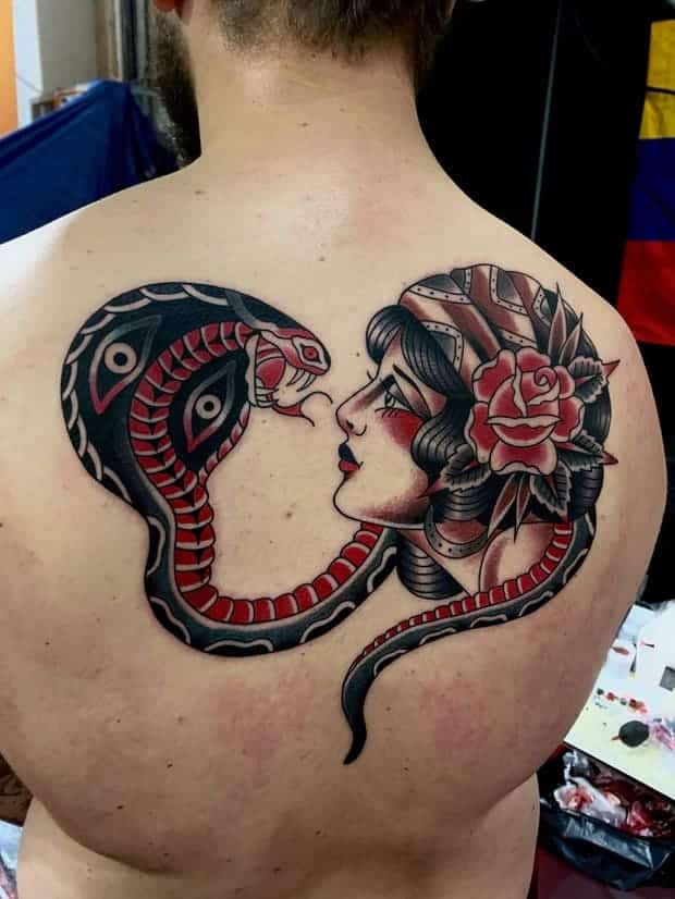 Traditional tattoo on the back of a man with a snake with an open mouth and woman with a flower in her hair looking at eachother.