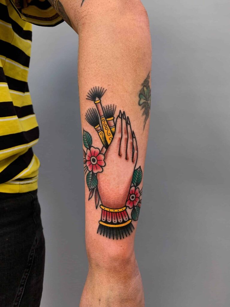 Traditional tattoo on a forearm of a hand holding three brushes with two flowers around it.