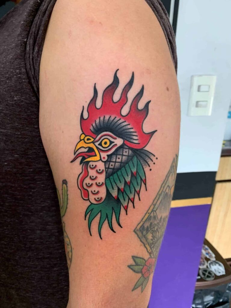 Traditional tattoo on a arm of a rooster with an open mouth and a tongue sticking out.