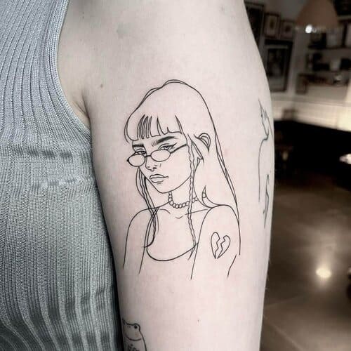 Fine line tattoo on a arm of a girl with glasses who looks upset and has a heartbroken symbol on her shoulder.